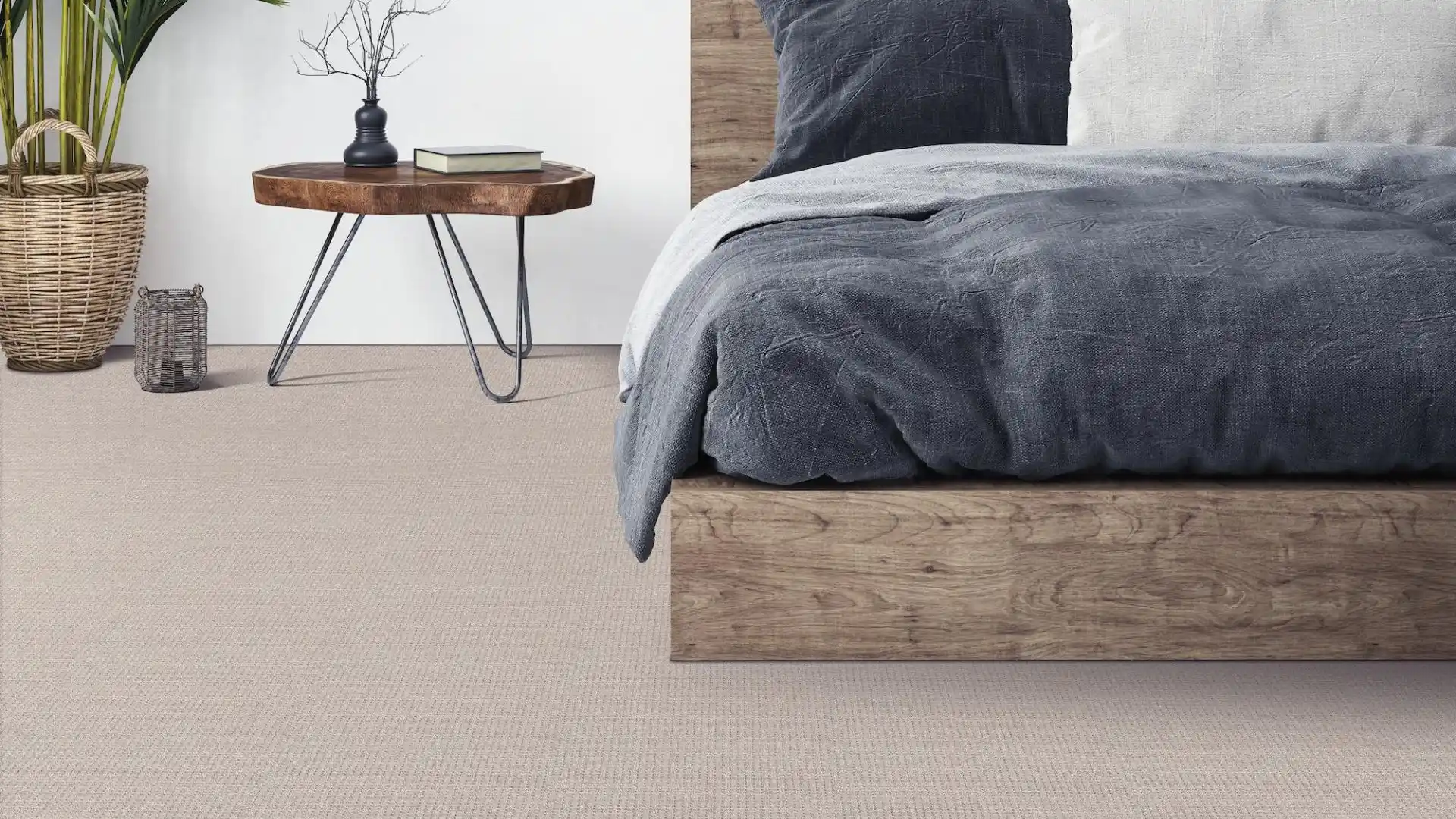 Carpet in bedroom, side of bed with blue covers, plant in corner, side wooden table.